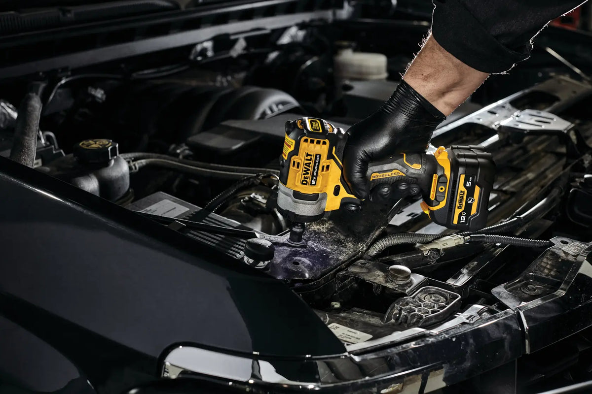 Close-up action shot of a worker using the DEWALT 12V MAX brushless cordless impact wrench to fasten components inside a car's engine bay, demonstrating precise speed control.