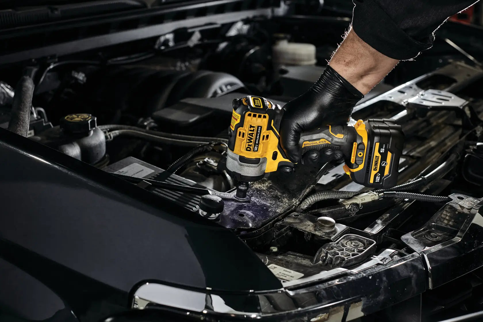 Close-up action shot of a worker using the DEWALT 12V MAX brushless cordless impact wrench to fasten components inside a car's engine bay, demonstrating precise speed control.