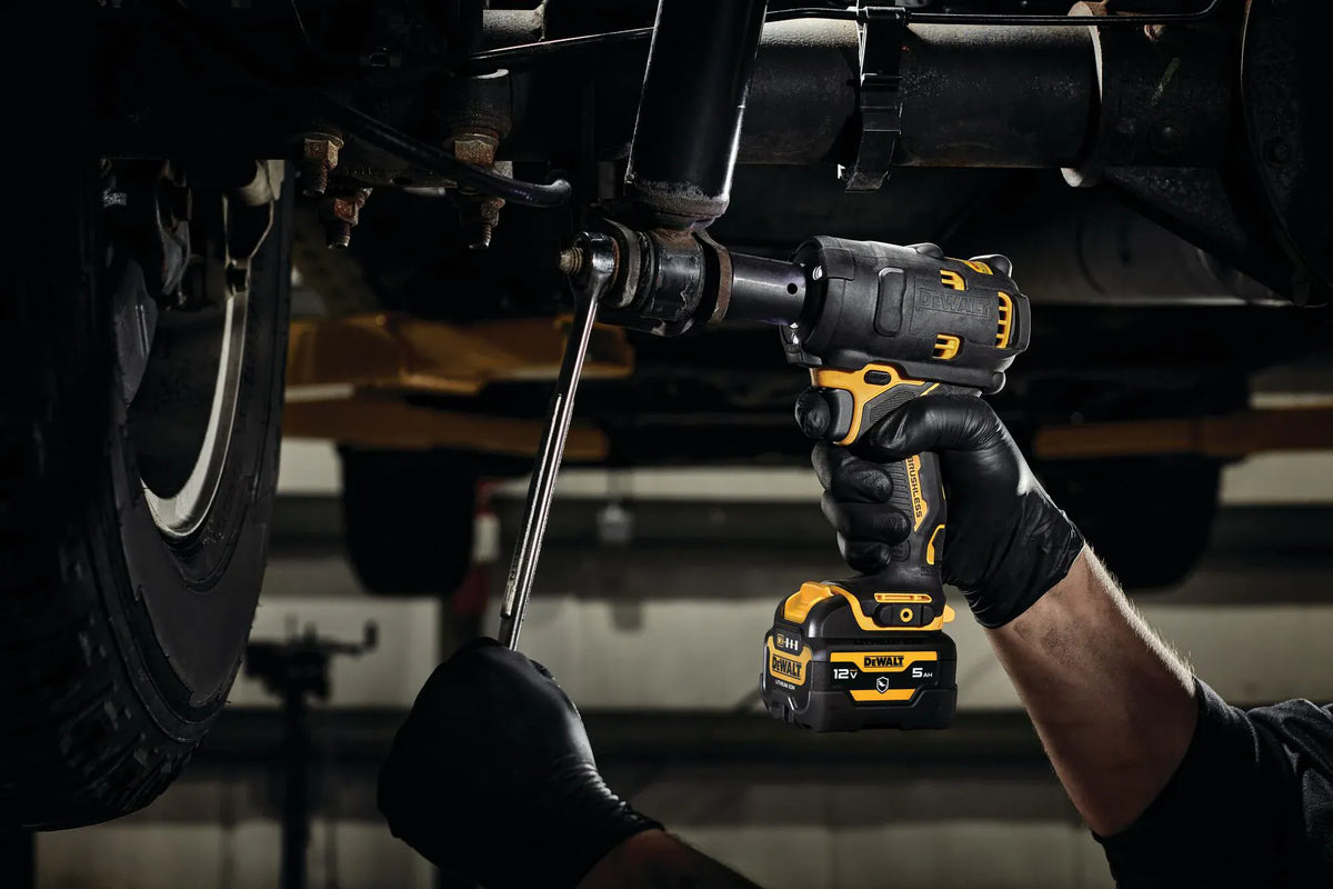Mechanic using the DEWALT DCF901B XTREME 12V MAX brushless 1/2-inch impact wrench with a 5.0Ah battery to remove a bolt from a vehicle's undercarriage in a dark shop environment.