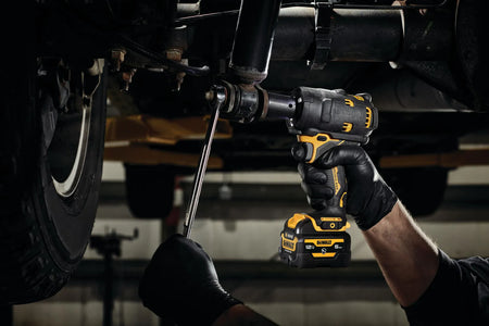 Mechanic using the DEWALT DCF901B XTREME 12V MAX brushless 1/2-inch impact wrench with a 5.0Ah battery to remove a bolt from a vehicle's undercarriage in a dark shop environment.