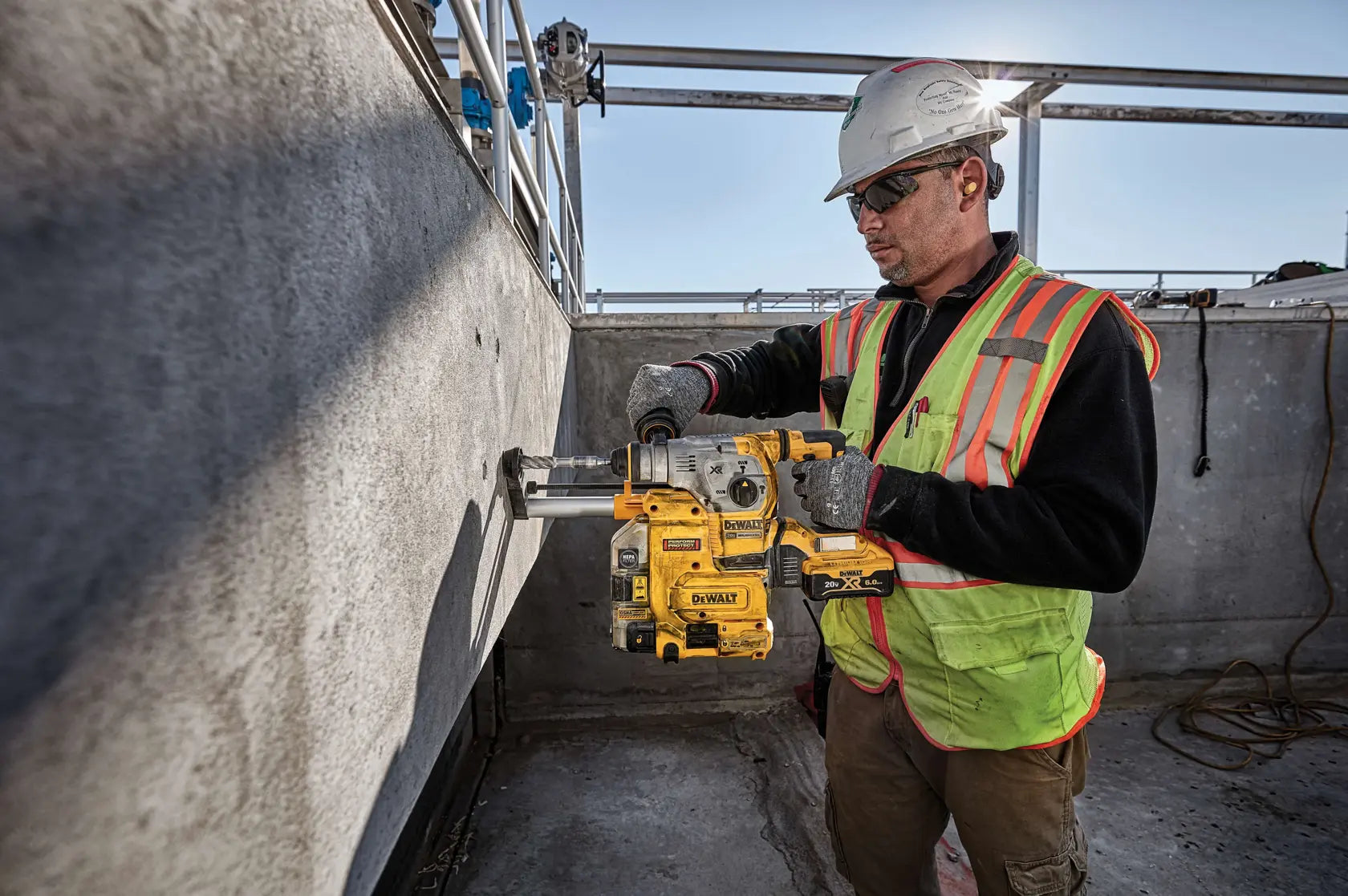 An action shot of a professional contractor using the DEWALT DCH293B rotary hammer to drill into a vertical concrete wall. The tool is shown equipped with an on-board dust extractor for OSHA Table 1 compliance, demonstrating its efficiency in industrial construction environments.