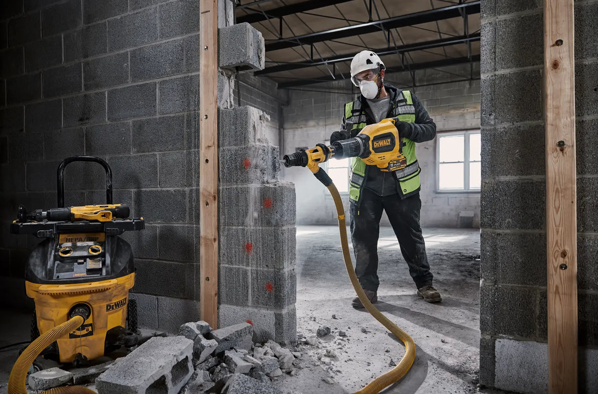 A professional contractor in full safety gear using the DEWALT DCH911 60V MAX chipping hammer with a dust extraction attachment to breach a concrete block wall in an industrial setting.