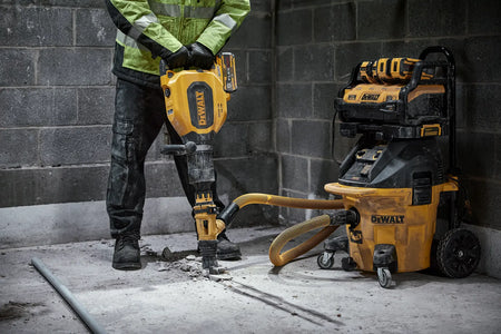 A construction professional using the DEWALT DCH911 60V MAX cordless chipping hammer to break up a concrete floor, featuring an attached dust extraction hose connected to a DEWALT HEPA vacuum for a clean and compliant demolition workspace.