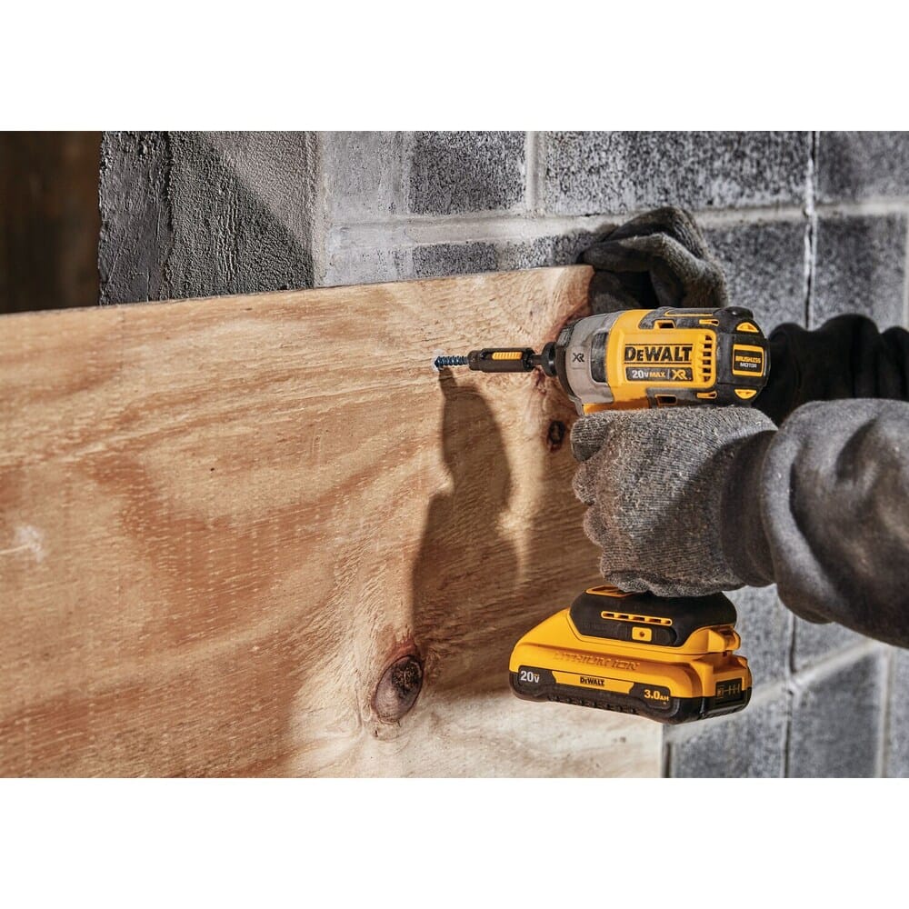 Action shot of a contractor using the DEWALT 20V MAX brushless impact driver to drive a fastener into a wood panel against a concrete block wall in a construction environment.