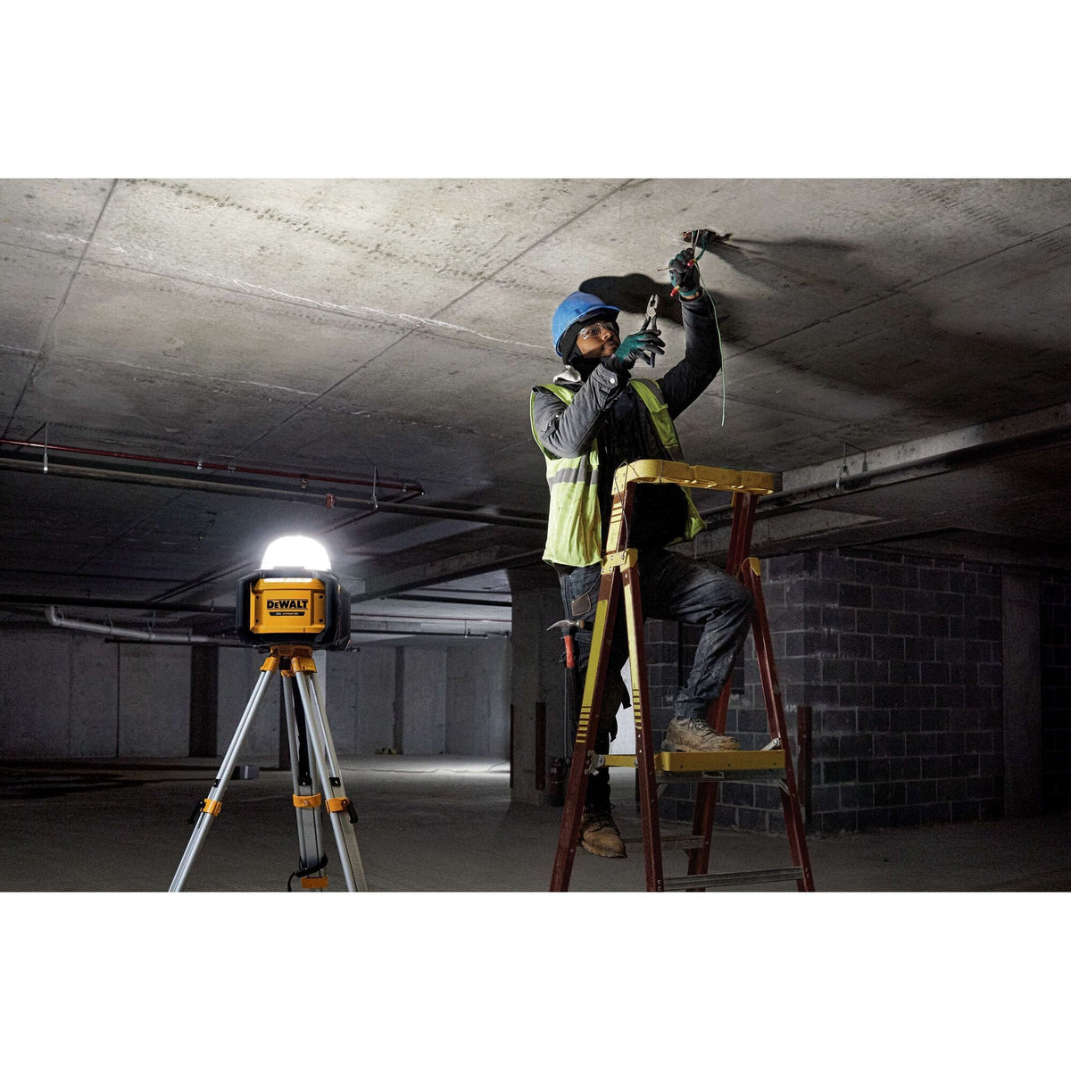 A professional electrician working on a ladder in a dark industrial basement, utilizing the DEWALT DCL074 5,000-lumen work light mounted on a tripod to provide high-intensity area illumination for safe wiring.