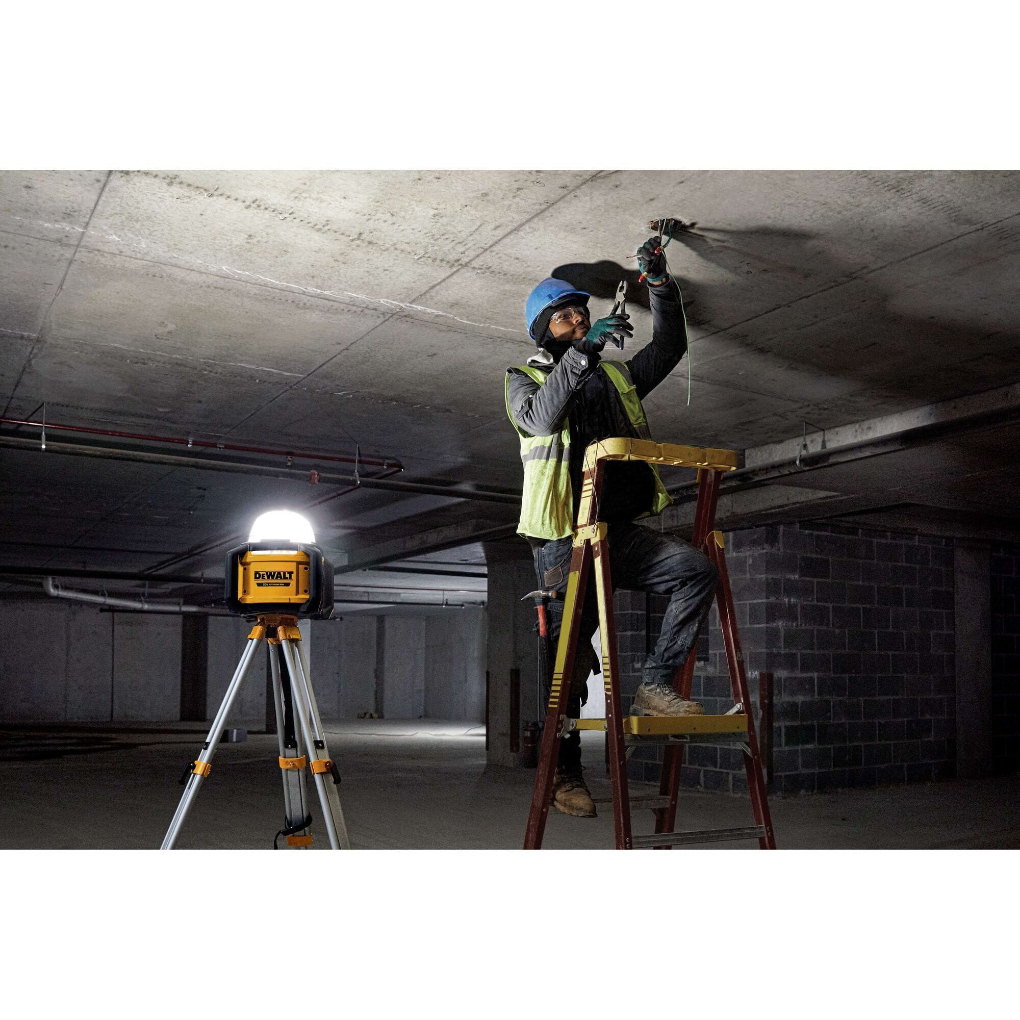 A professional electrician working on a ladder in a dark industrial basement, utilizing the DEWALT DCL074 5,000-lumen work light mounted on a tripod to provide high-intensity area illumination for safe wiring.