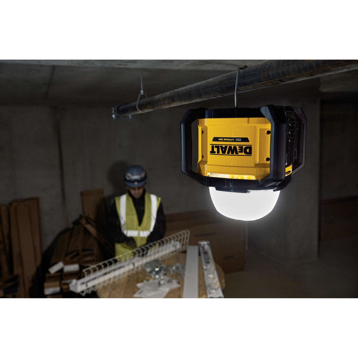 Wide action shot of the DEWALT 20V MAX cordless area light hanging from a conduit pipe, providing powerful 360-degree overhead lighting for a professional tradesperson organizing materials in a dim workspace.