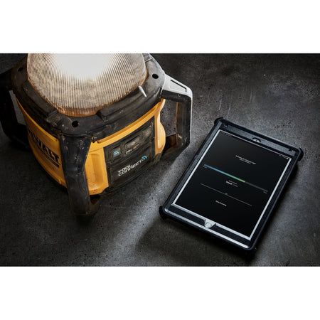 Close-up shot of a tablet displaying the DEWALT Tool Connect dashboard next to the DCL074 LED light, highlighting the ability to monitor battery life and signal strength on the jobsite.