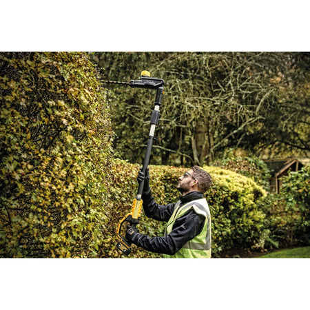 A technician utilizing the articulating head of the DEWALT cordless pole trimmer to vertically square off the side of a tall privacy hedge, demonstrating the tool's 12-foot maximum reach.