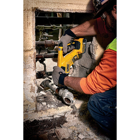 A tradesman using the DEWALT 20V MAX XR Compact Reciprocating Saw to cut a thick metal pipe inside a tight concrete wall opening, showcasing the tool's 14.5 inch compact length and effectiveness for demolition work in confined spaces.