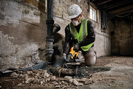 Action shot showing a professional plumber in a basement using the DEWALT 60V MAX reciprocating saw to cut through thick iron waste pipes. The technician is wearing full safety gear, demonstrating the tool's cordless maneuverability and high material removal rate in cramped environments.