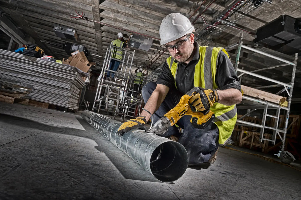 A professional tradesperson wearing a hard hat and safety gear kneeling on a job site while using the DEWALT 20V MAX cordless metal shears to precisely cut a large galvanized steel ventilation duct.