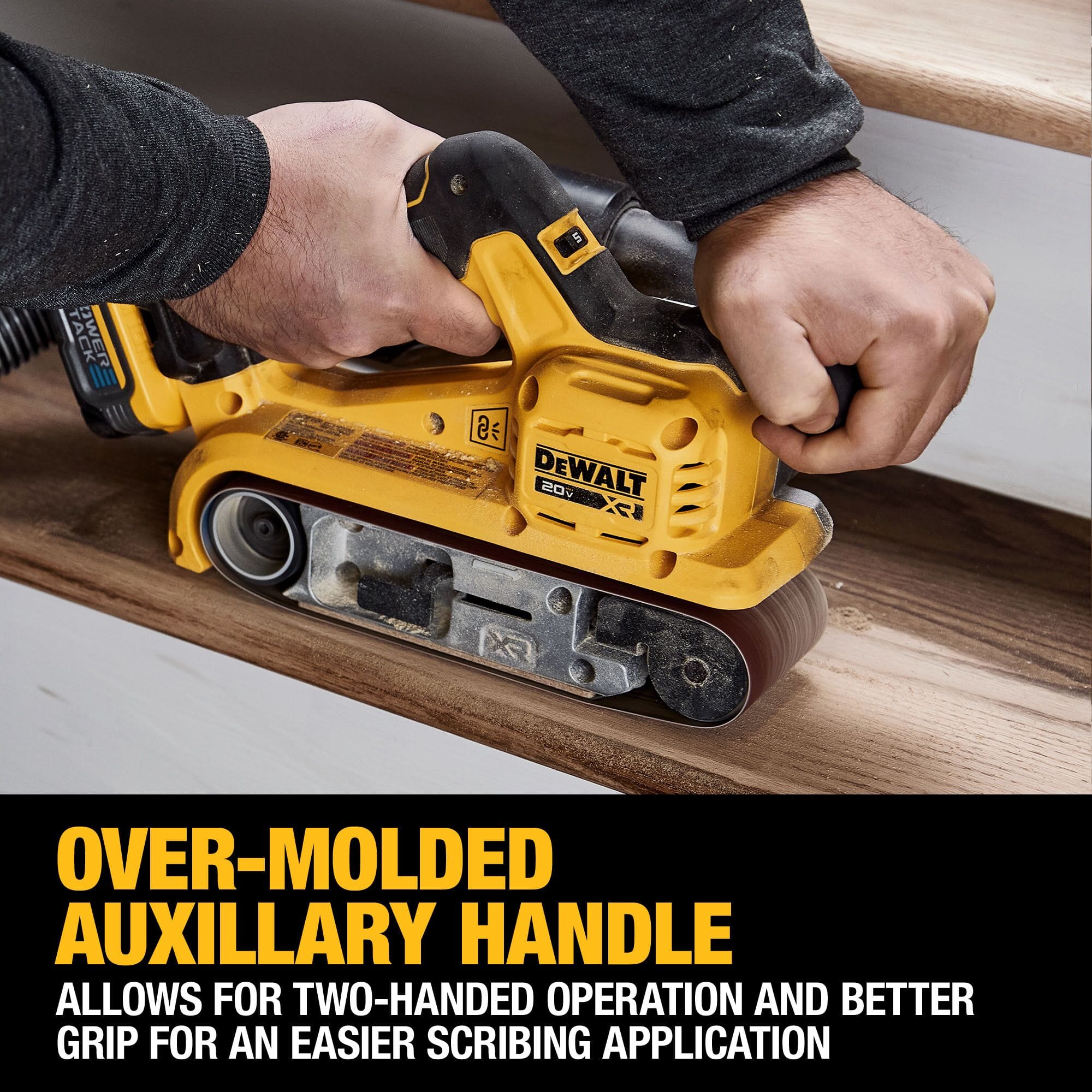 Action shot highlighting the ergonomics of the DEWALT cordless belt sander. A user is shown gripping the over-molded auxiliary handle for precise two-handed operation, with text noting this design provides a better grip for easier scribing and aggressive material removal applications.