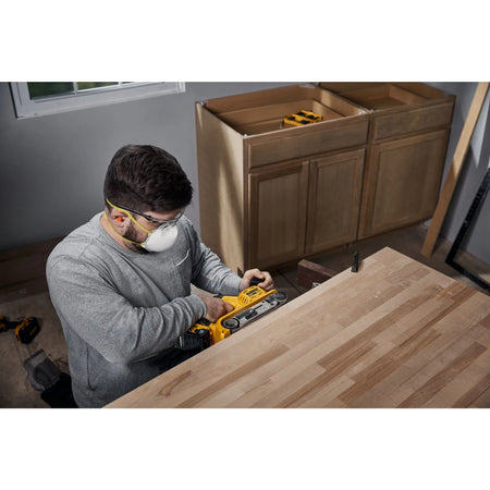Action shot of a professional woodworker in a workshop using the DEWALT belt sander to level a large tabletop. The image showcases the cordless freedom and the use of a dust mask and safety glasses, emphasizing professional jobsite safety and productivity.