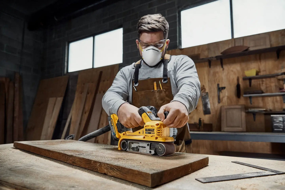 Action shot of a craftsman using the DCW222B belt sander to finish a thick wooden slab. The technician is using both hands for maximum control while the integrated vacuum hose connection keeps the workpiece clear of debris for better visibility.