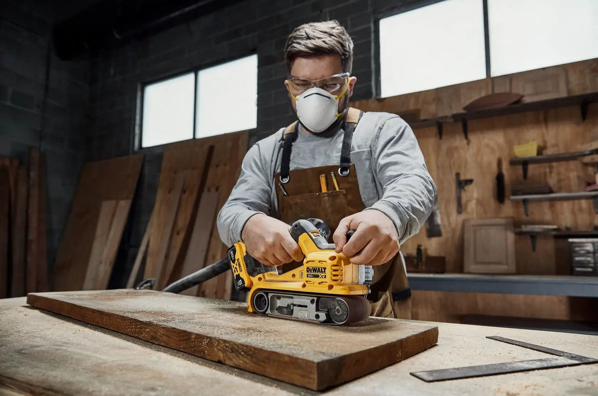 Front-facing action shot of a professional woodworker operating the DEWALT 20V MAX XR belt sander. This view highlights the auxiliary handle placement and the aggressive material removal capabilities of the 3x21-inch belt on a hardwood surface.