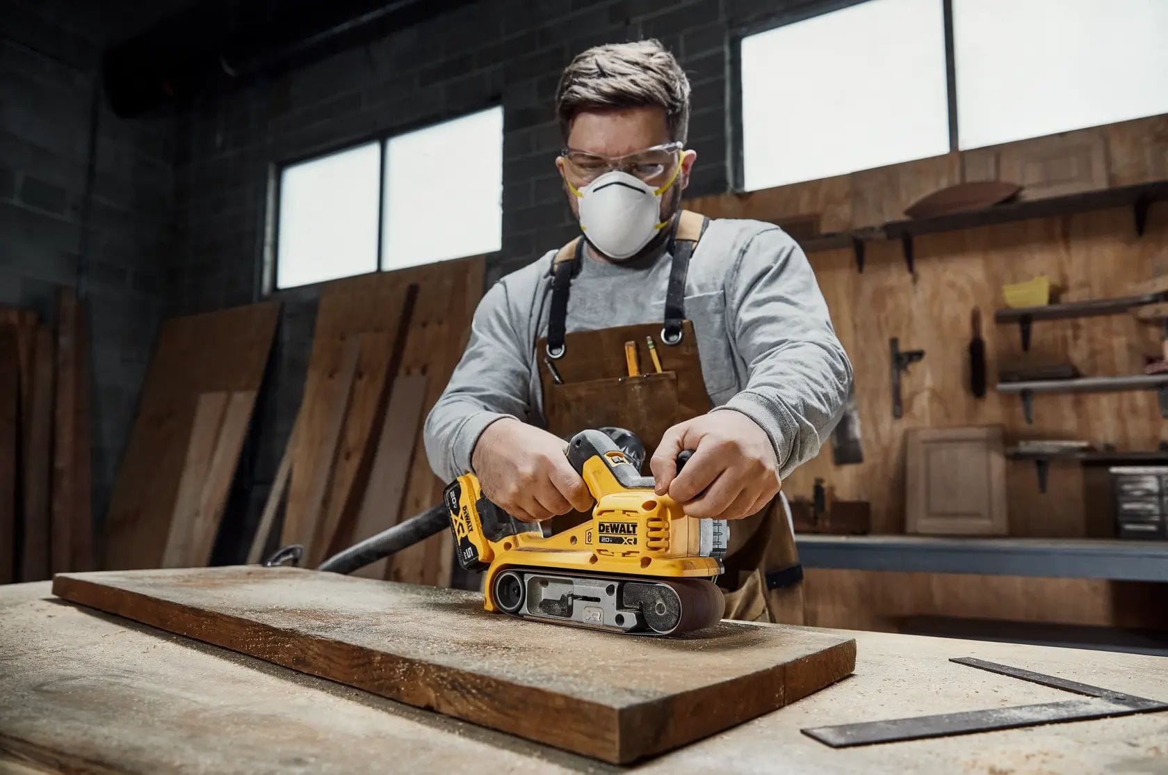 Front-facing action shot of a professional woodworker operating the DEWALT 20V MAX XR belt sander. This view highlights the auxiliary handle placement and the aggressive material removal capabilities of the 3x21-inch belt on a hardwood surface.