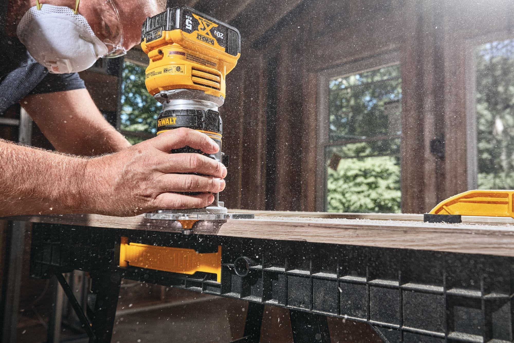 A carpenter wearing a protective dust mask operates the DEWALT 20V MAX XR cordless compact router to machine a precise profile along the edge of a long hardwood board on a jobsite workbench.