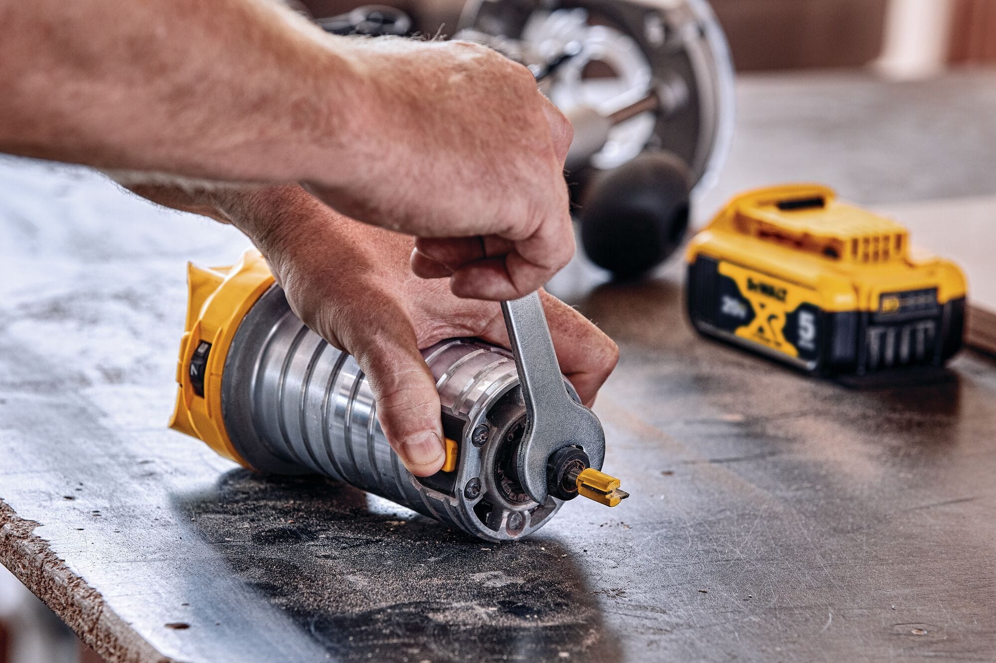 A close-up view of a woodworker's hands using a flat steel collet wrench to secure a yellow-fluted router bit into the 1/4-inch steel chuck of the detached router motor housing.