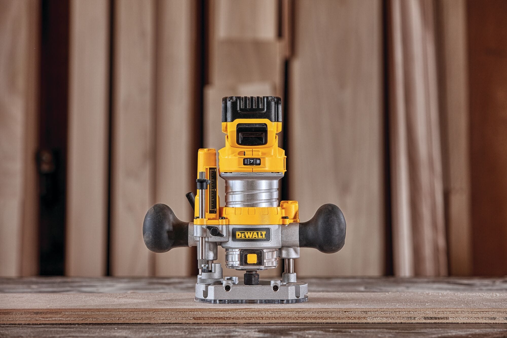 The DEWALT compact router motor unit is shown mounted inside an adjustable aluminum plunge base accessory on a wooden board, highlighting the dual rubber side handles and vertical depth adjustment rod.
