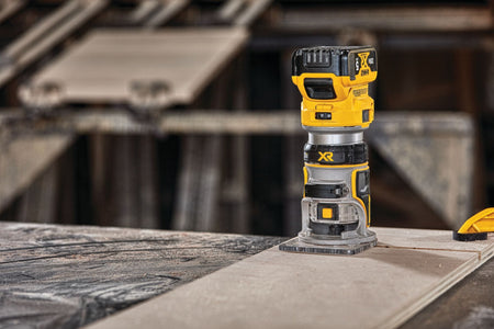 The DEWALT cordless compact fixed-base router stands upright on a large sheet of plywood material inside an active woodworking workshop, ready for wood edge trimming applications.