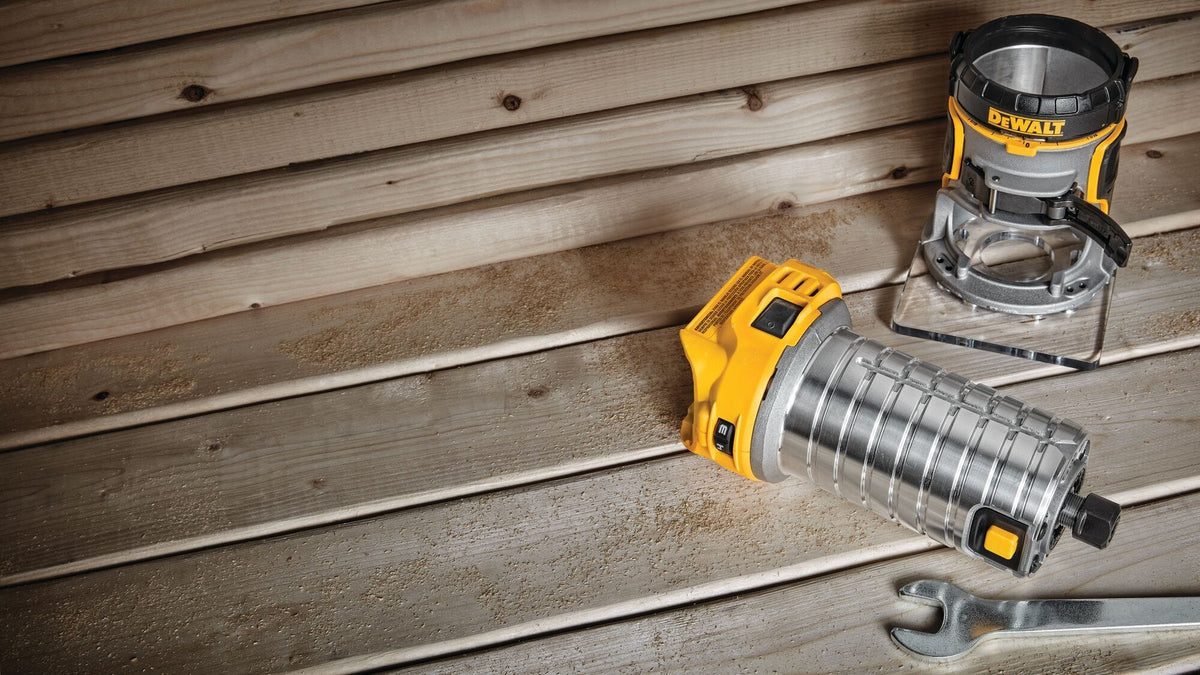 The detached aluminum cylindrical motor of the DEWALT compact router lies horizontally on a wooden board surface next to its empty fixed base platform and an open-ended adjustment wrench.