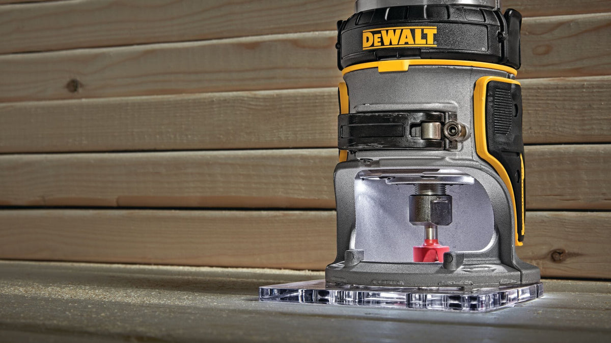 A low-angle macro view of the clear acrylic sub-base on the DEWALT compact router, showing the integrated dual LEDs brightly illuminating a red carbide-tipped routing bit as it shapes a wood plank.