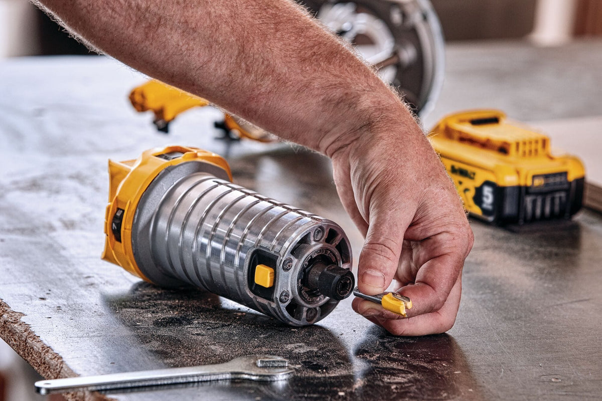 A close-up detailed shot of a tradesman's hands inserting a yellow-fluted wood profiling router bit directly into the open 1/4-inch steel collet chuck of the detached router motor cylinder lying on a workbench table.