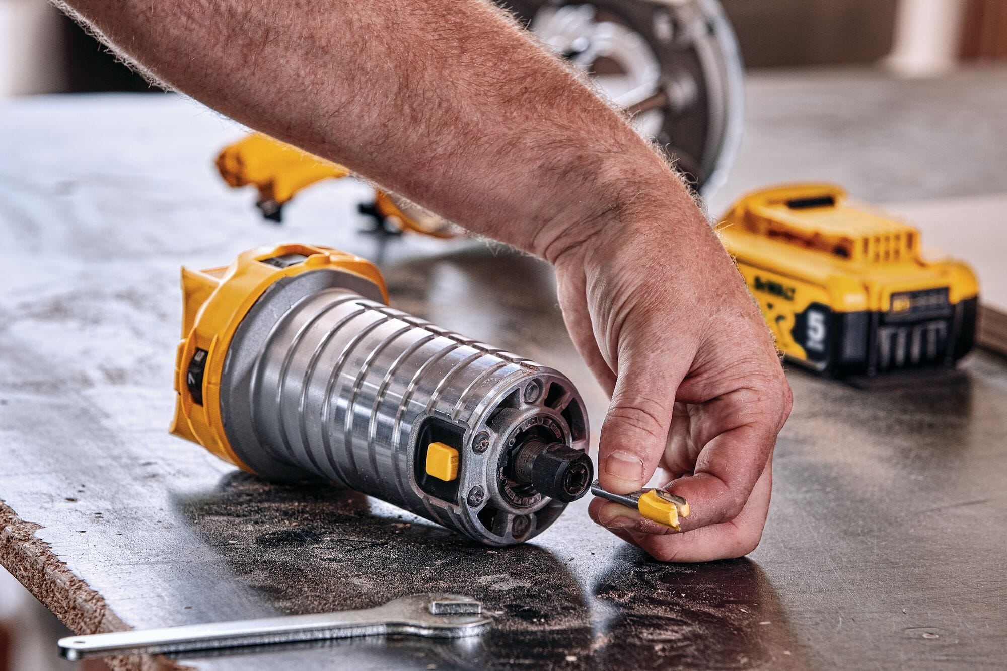 A close-up detailed shot of a tradesman's hands inserting a yellow-fluted wood profiling router bit directly into the open 1/4-inch steel collet chuck of the detached router motor cylinder lying on a workbench table.