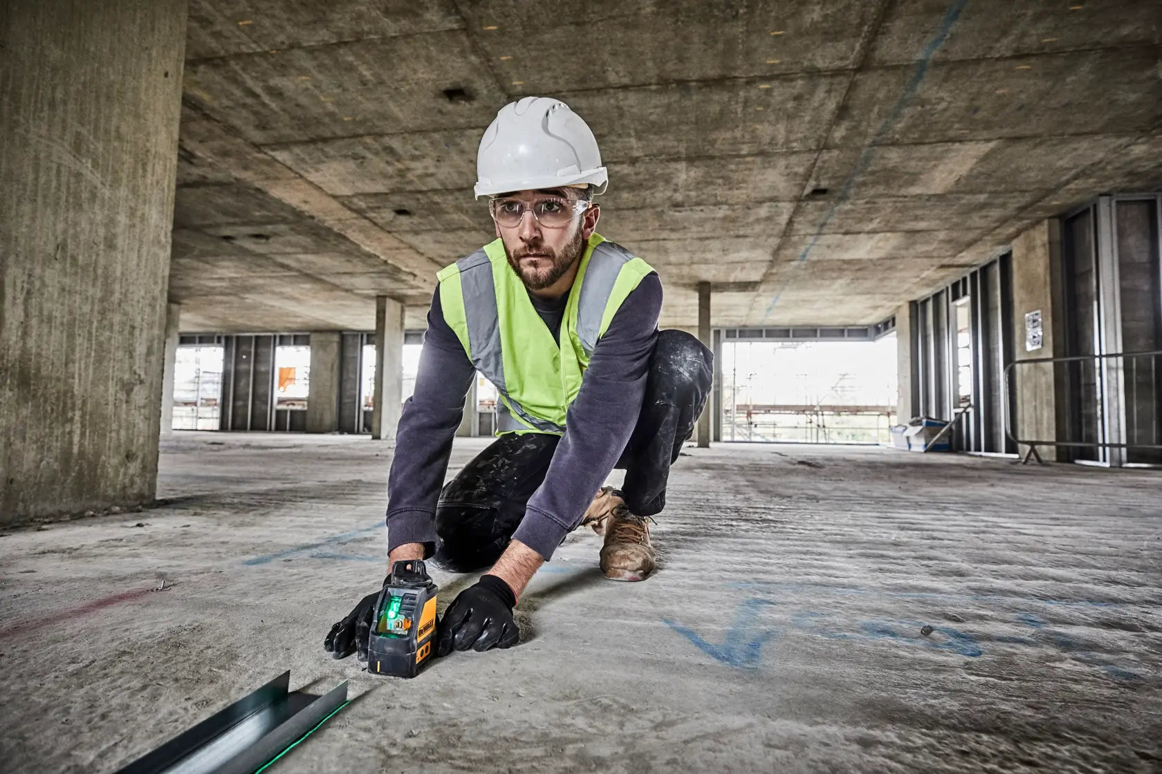 A professional contractor wearing a hard hat and safety vest kneeling on a concrete floor while using the DEWALT green cross line laser to align metal floor tracks for wall framing.