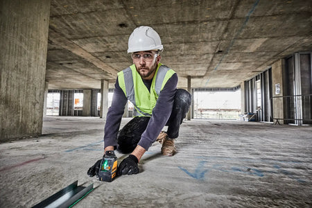 A professional contractor wearing a hard hat and safety vest kneeling on a concrete floor while using the DEWALT green cross line laser to align metal floor tracks for wall framing.
