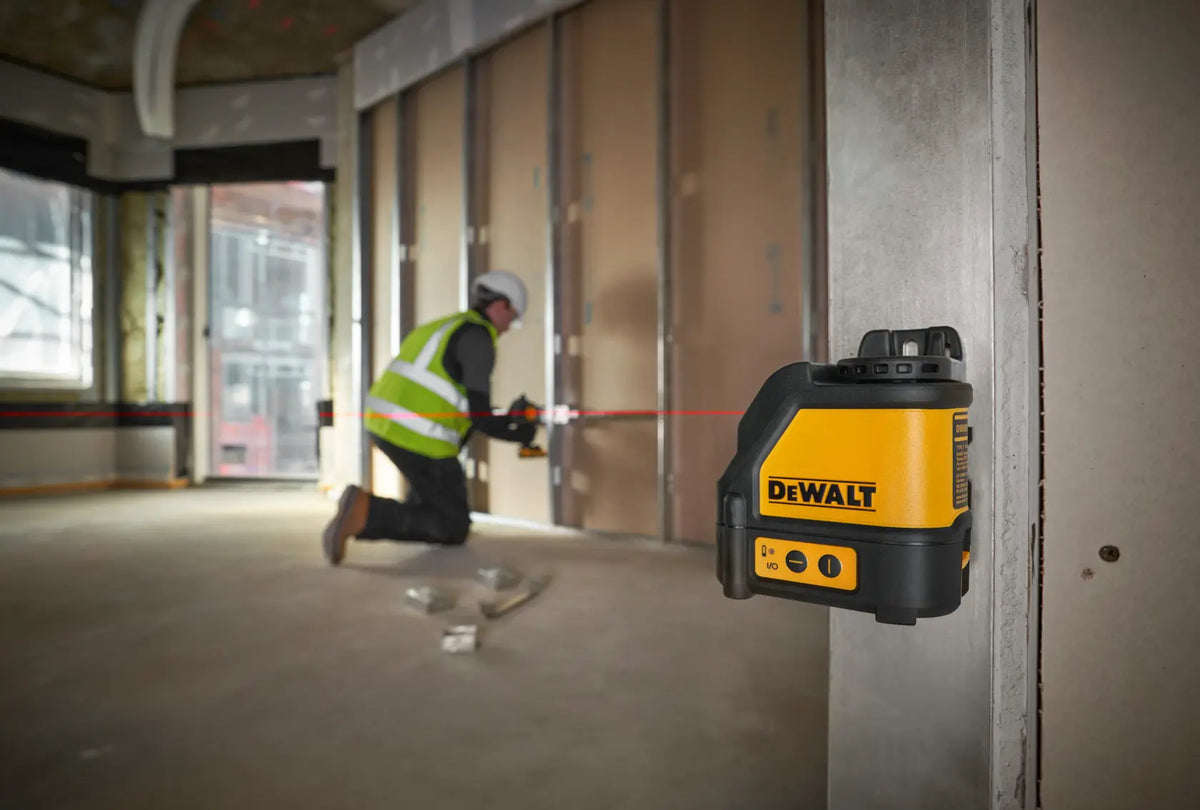 An action lifestyle shot showing the DEWALT DW088K magnetic laser level mounted to a steel track in a commercial building. A professional contractor is visible in the background using the projected red horizontal line for precise alignment of electrical boxes and wall framing.