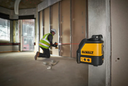 An action lifestyle shot showing the DEWALT DW088K magnetic laser level mounted to a steel track in a commercial building. A professional contractor is visible in the background using the projected red horizontal line for precise alignment of electrical boxes and wall framing.