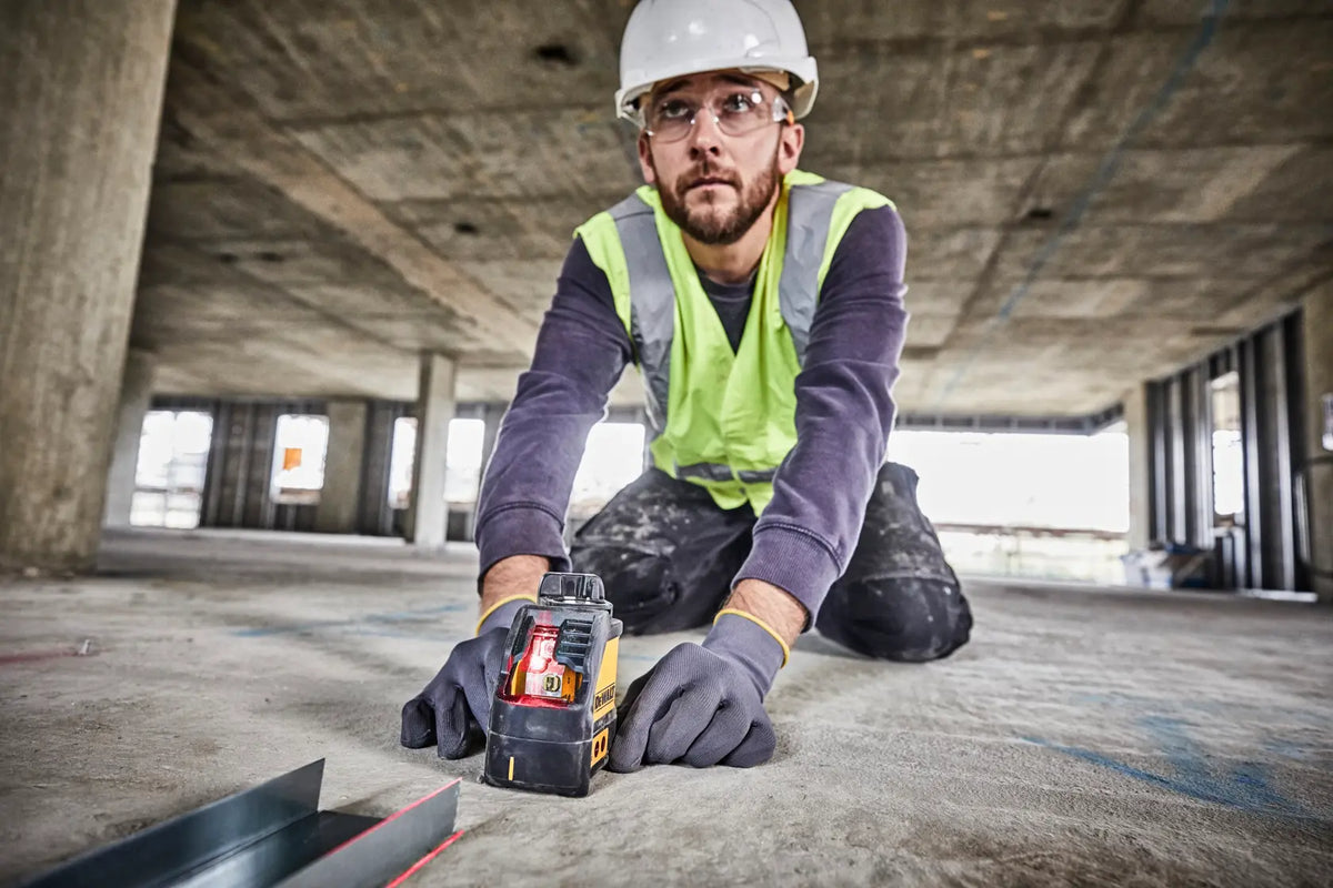 An action lifestyle shot of a professional installer wearing a hard hat and safety glasses kneeling behind the DEWALT self-leveling laser. The user is checking the alignment of a metal floor track against the bright red vertical laser beam in a large-scale industrial workspace.