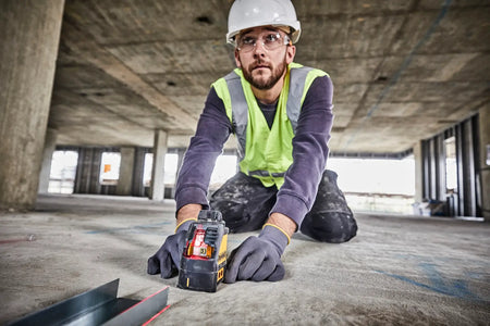 An action lifestyle shot of a professional installer wearing a hard hat and safety glasses kneeling behind the DEWALT self-leveling laser. The user is checking the alignment of a metal floor track against the bright red vertical laser beam in a large-scale industrial workspace.