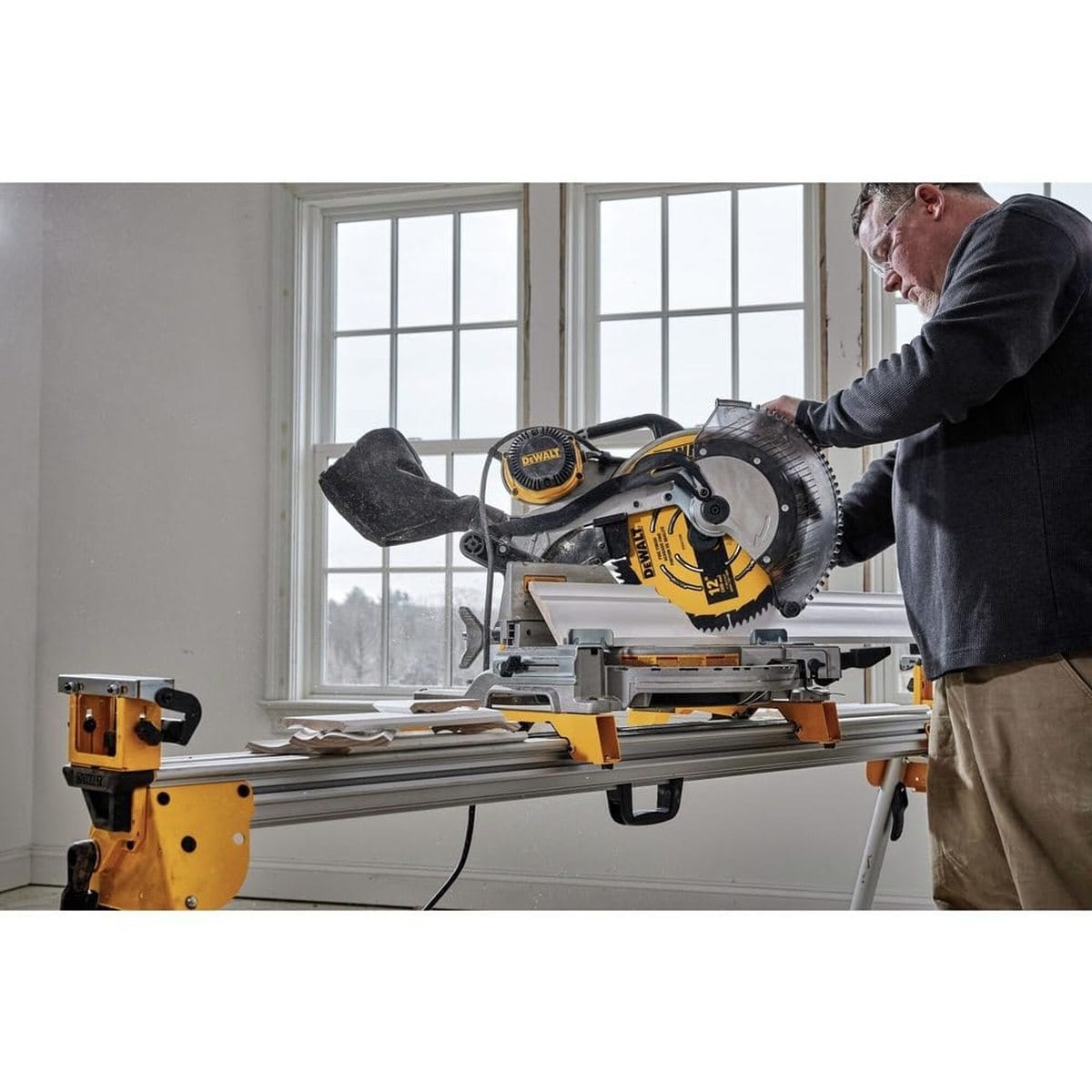 A professional woodworker using a DEWALT sliding miter saw equipped with a 12-inch carbide blade to make a precise crosscut through white crown molding in a residential setting.