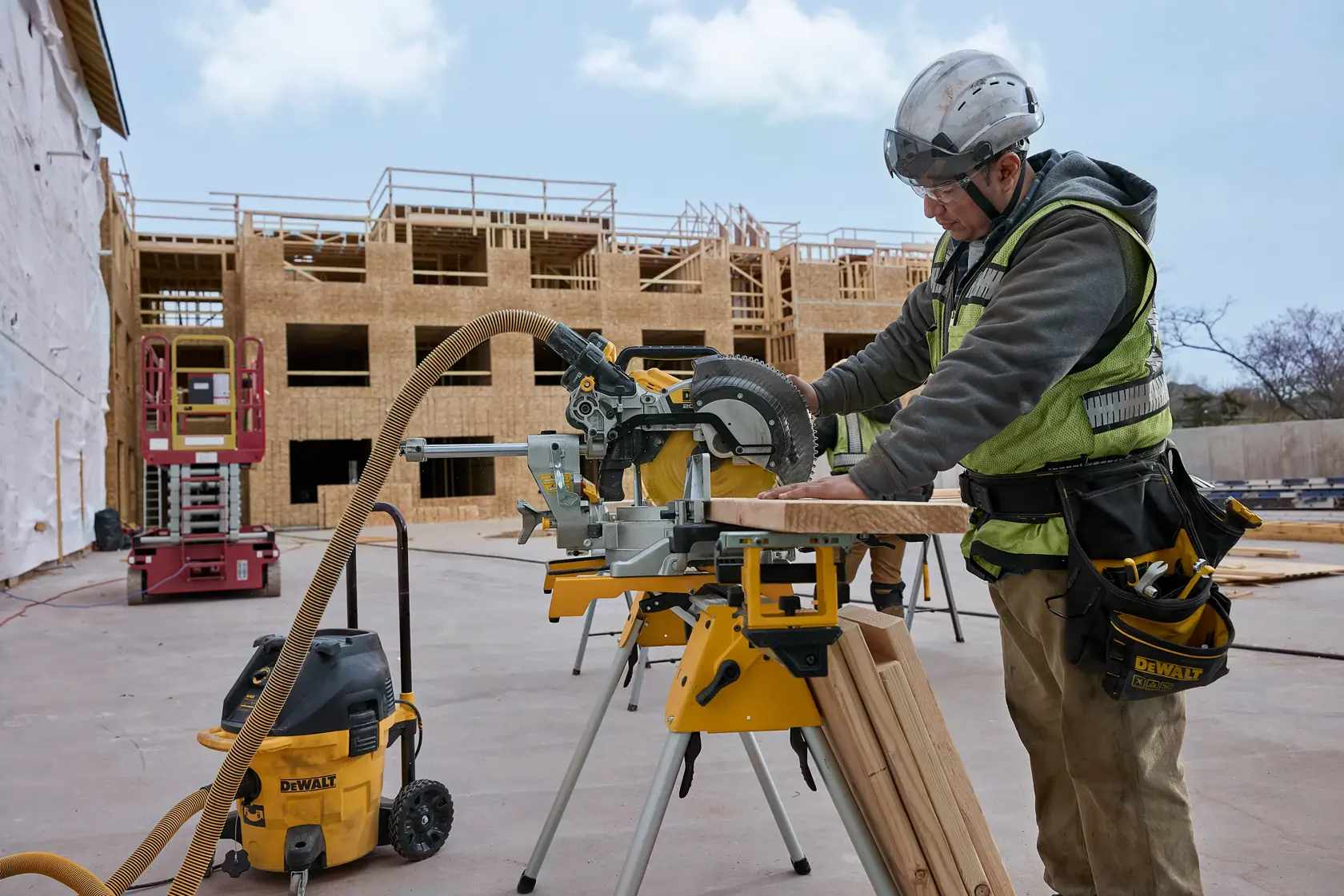 Jobsite application photo showing a worker using a DEWALT miter saw and 12-inch carbide blade kit integrated with a dust extraction vacuum system to maintain a clean workspace during framing.