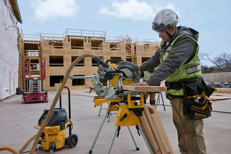 Jobsite application photo showing a worker using a DEWALT miter saw and 12-inch carbide blade kit integrated with a dust extraction vacuum system to maintain a clean workspace during framing.