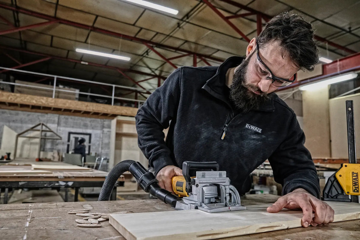 Professional woodworker wearing safety glasses using the DEWALT DW682K 6.5 Amp corded electric biscuit plate joiner with an attached vacuum hose for clean, heavy-duty carpentry work.
