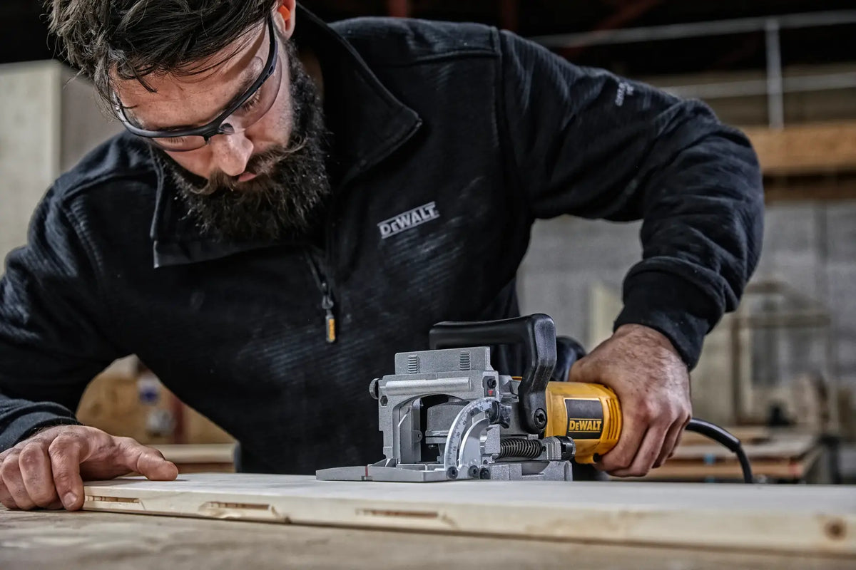 Carpenter utilizing the DEWALT DW682K heavy-duty woodworking biscuit joiner power tool to plunge a precise slot into the edge of a wooden board.