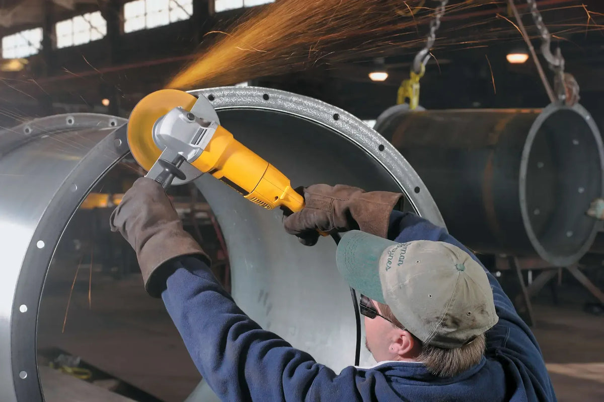 A wide lifestyle shot of a tradesman in protective gear using the DEWALT 7-inch angle grinder to smooth the edges of a large industrial steel pipe. The image illustrates the tool's stability and power when used for heavy-duty metal fabrication and pipe fitting.