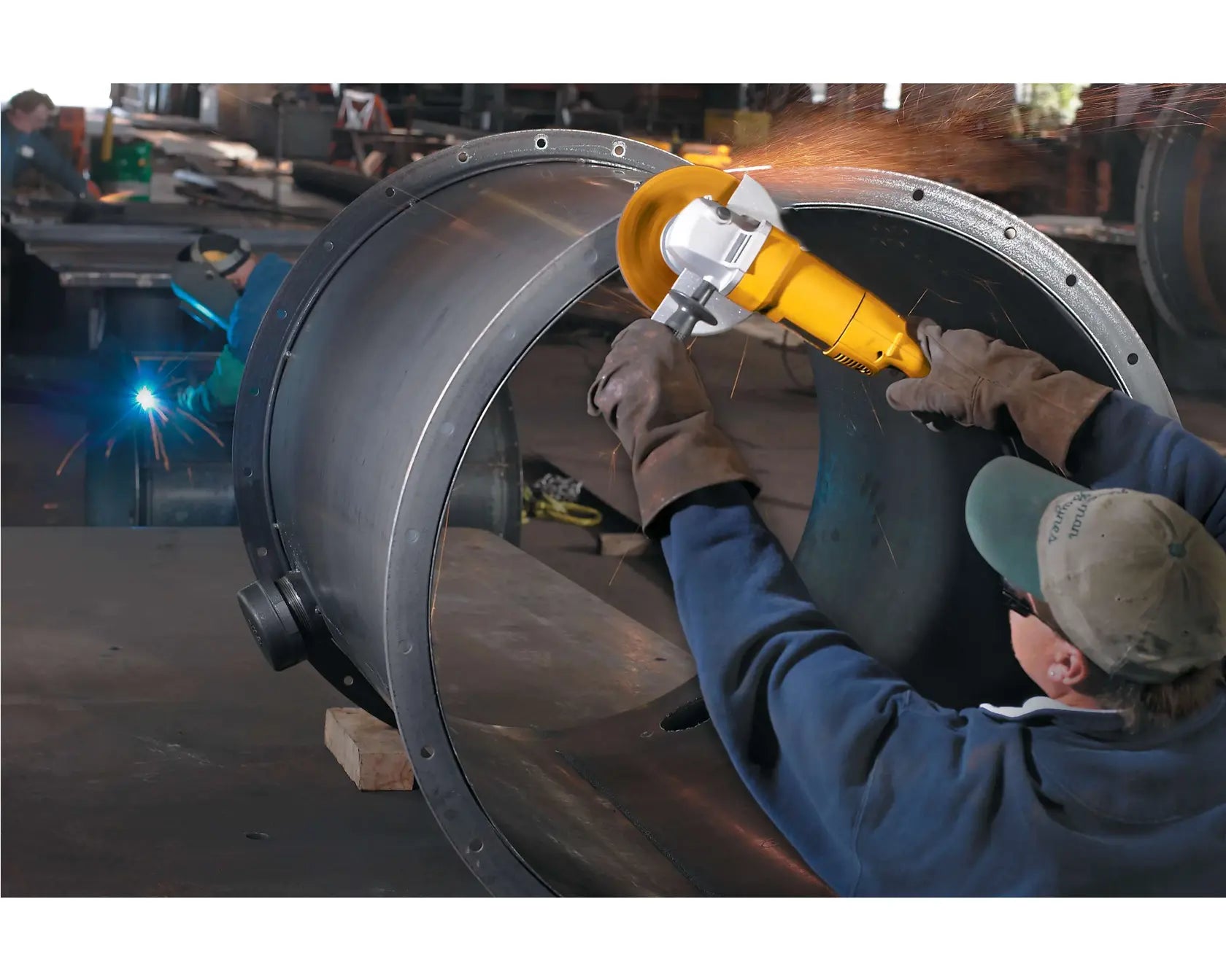 A wide action lifestyle shot of a professional welder using the DEWALT DW840 7-inch angle grinder to prep the surface of a large industrial steel cylinder. The 13-amp corded motor provides high-torque power for heavy-duty metal fabrication while the adjustable guard redirects sparks safely away from the operator.