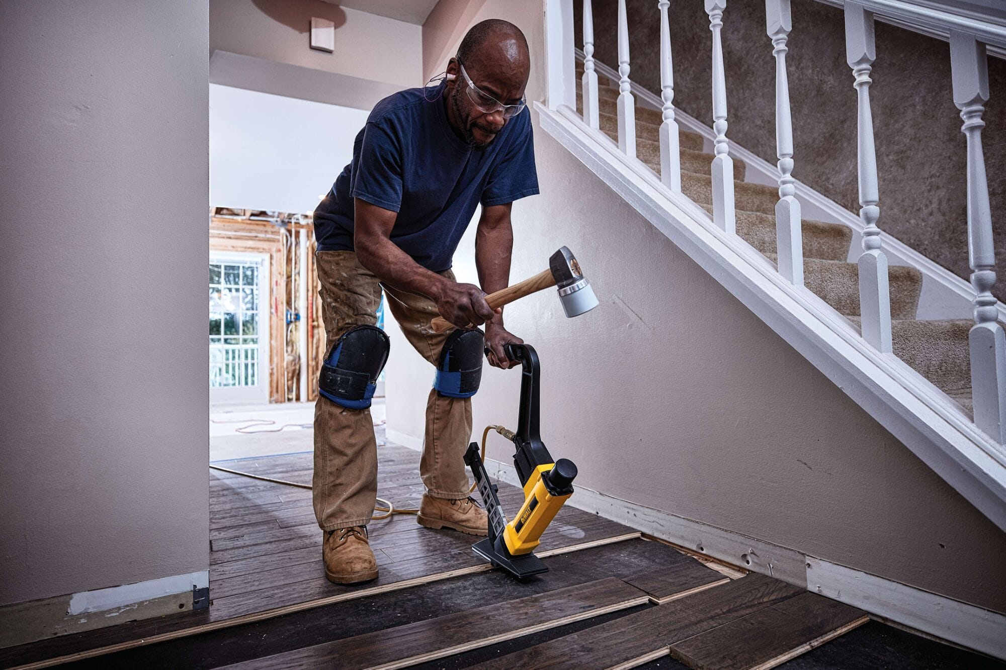 A tradesperson using the DEWALT 2-in-1 flooring tool and mallet to secure engineered wood flooring near a staircase, illustrating the tool's maneuverability and professional results in residential construction.