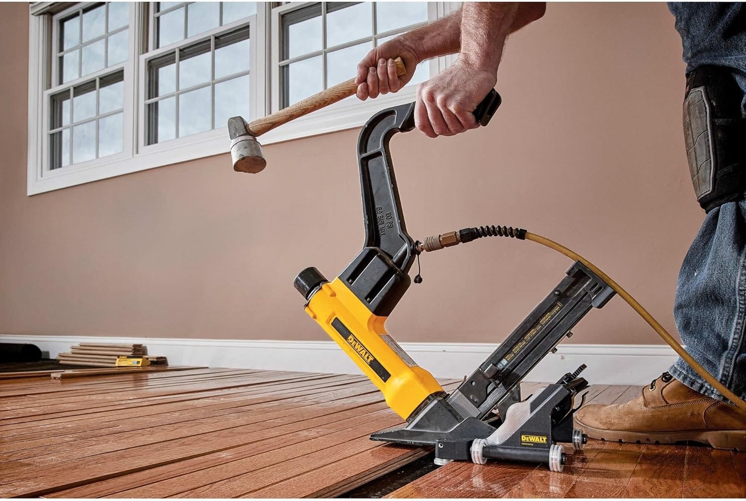 Action shot of a professional installer using a mallet to strike the actuator of the DEWALT pneumatic flooring tool, featuring the integrated rolling base attachment for rapid and consistent fastening of hardwood planks.