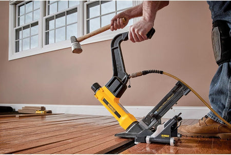 Action shot of a professional installer using a mallet to strike the actuator of the DEWALT pneumatic flooring tool, featuring the integrated rolling base attachment for rapid and consistent fastening of hardwood planks.