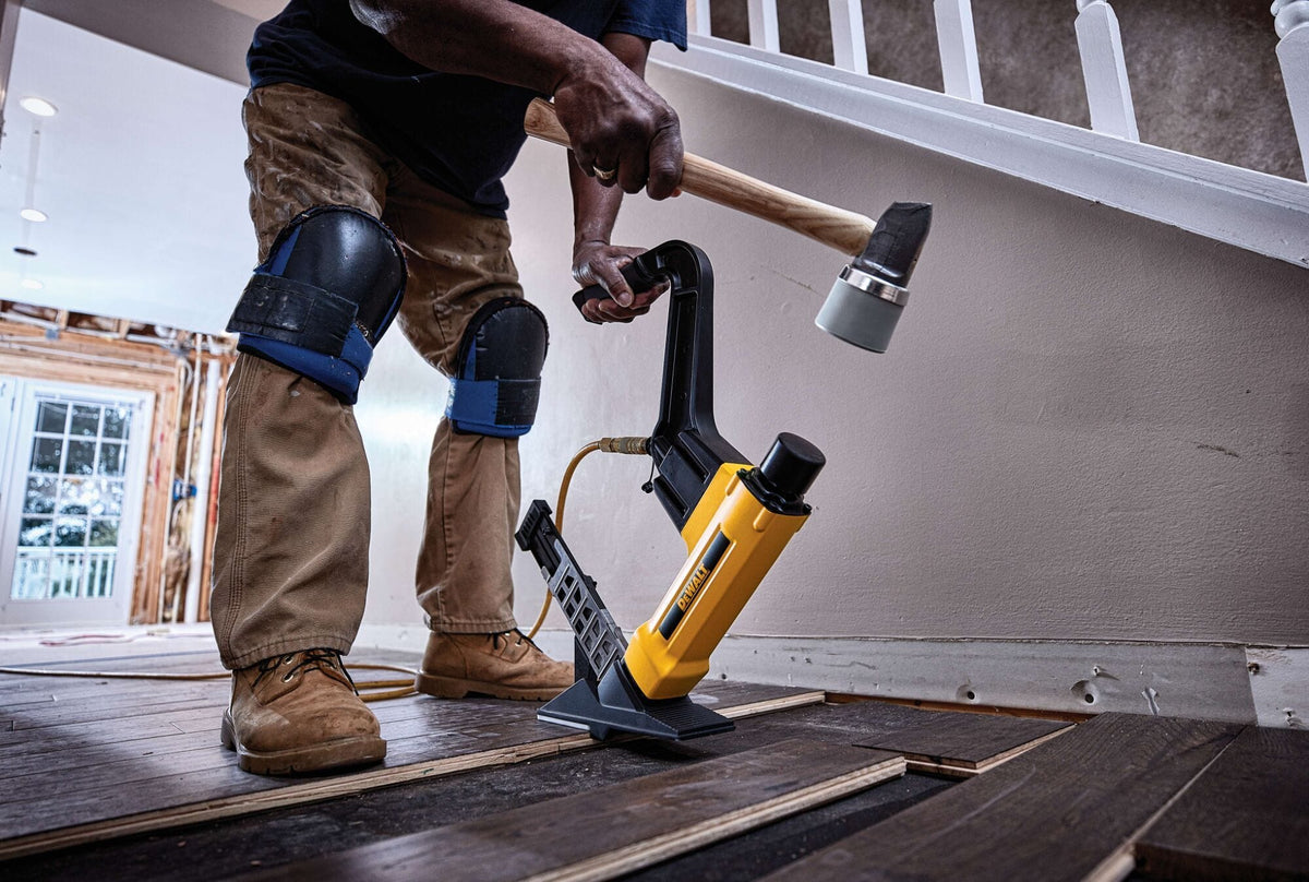 A professional flooring contractor wearing knee pads using the DEWALT DWFP12569 nailer and a mallet to install dark hardwood planks, demonstrating the tool's ergonomic reach and pneumatic driving power.