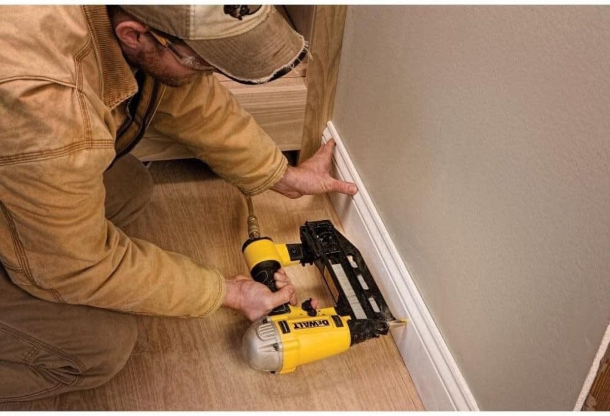 Installer fastening a white baseboard using the DEWALT DWFP71917 pneumatic 16‑gauge finish nailer, showing controlled placement, ergonomic handling, and clean finish fastening ideal for trim carpentry and interior molding installation