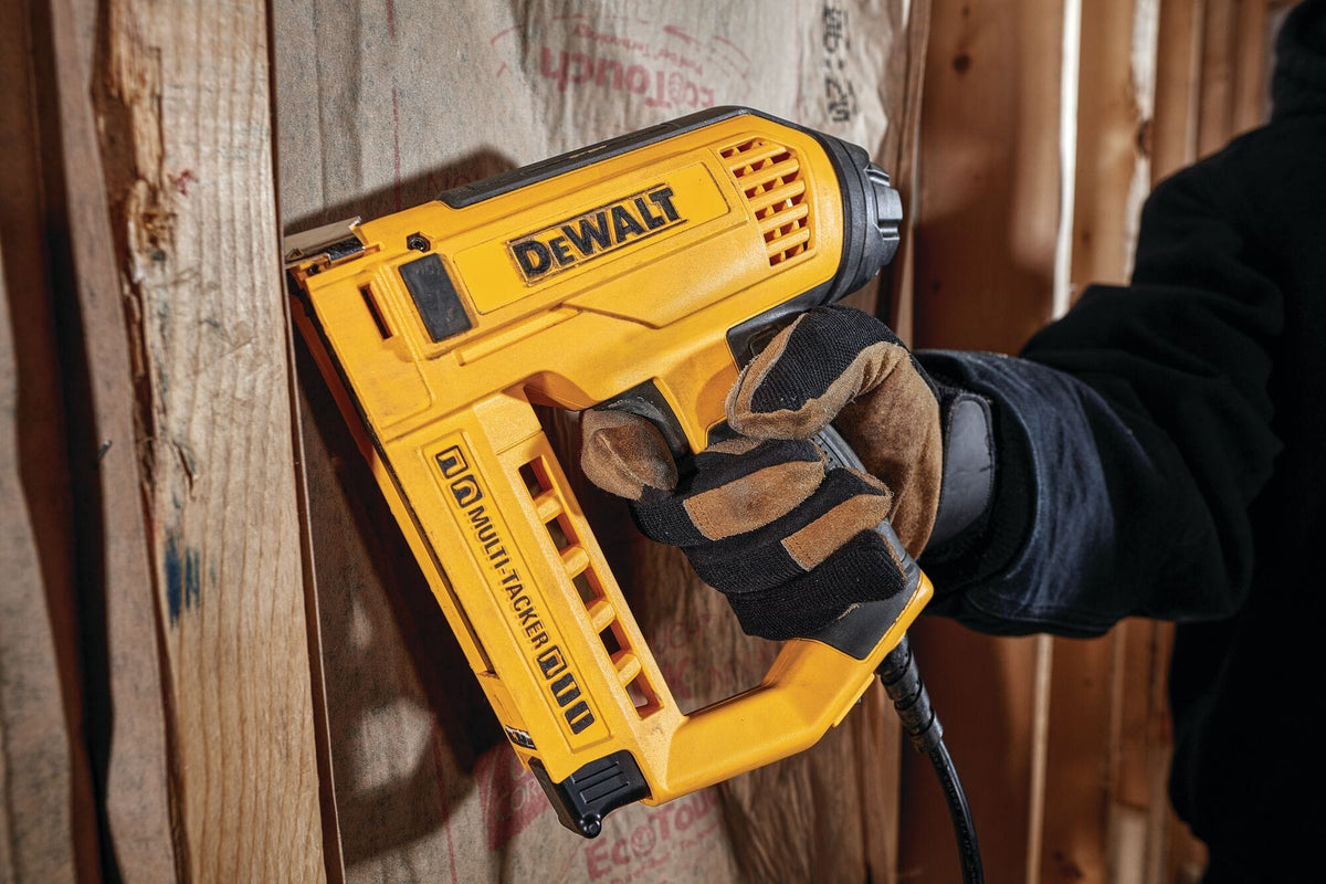 A worker wearing protective gloves uses the DEWALT 5-in-1 Electric Tacker to secure insulation or underlayment to a wooden stud, demonstrating the tool's compact size and one-handed operation.