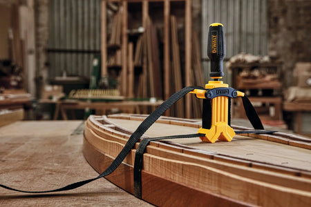 An action shot of the DEWALT DWHT83839 band clamp being used in a professional woodworking shop. The black nylon strap is shown tightly wrapped around a curved, irregular wooden workpiece on a workbench, illustrating how the 1,000 lbs of clamping force secures complex shapes during glue-up.