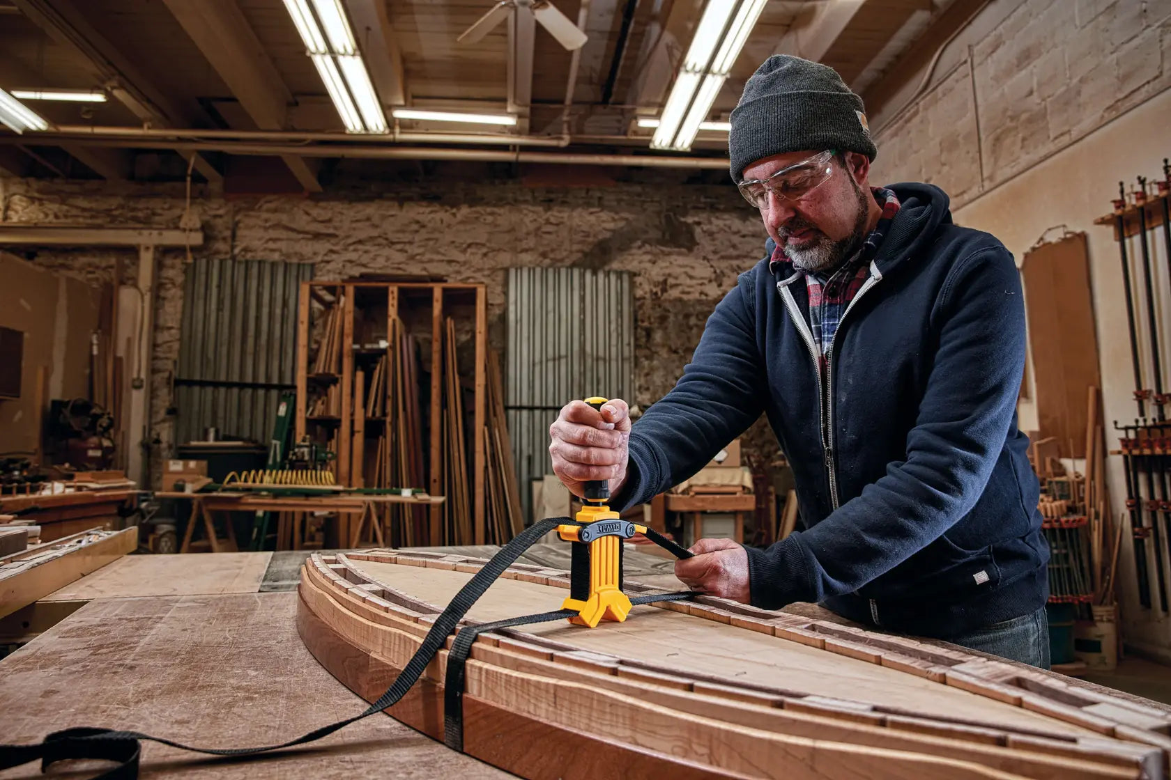 A lifestyle shot featuring a craftsman using the DEWALT heavy-duty band clamp to secure a wooden frame. The technician is shown turning the ergonomic quad-lobe handle to apply high-tension torque, demonstrating the tool's effectiveness for DIY furniture repair and professional cabinetry tasks.